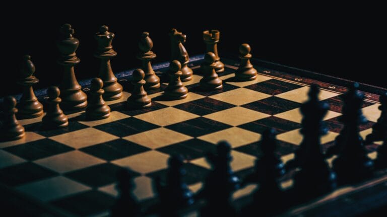 لقطة قريبة لقطع شطرنج خشبية في موضع بداية اللعبة على رقعة شطرنج خشبية تقليدية، بإضاءة درامية منخفضة تبرز عمق استراتيجية الشطرنج والتخطيط. دليل تعلم الشطرنج.