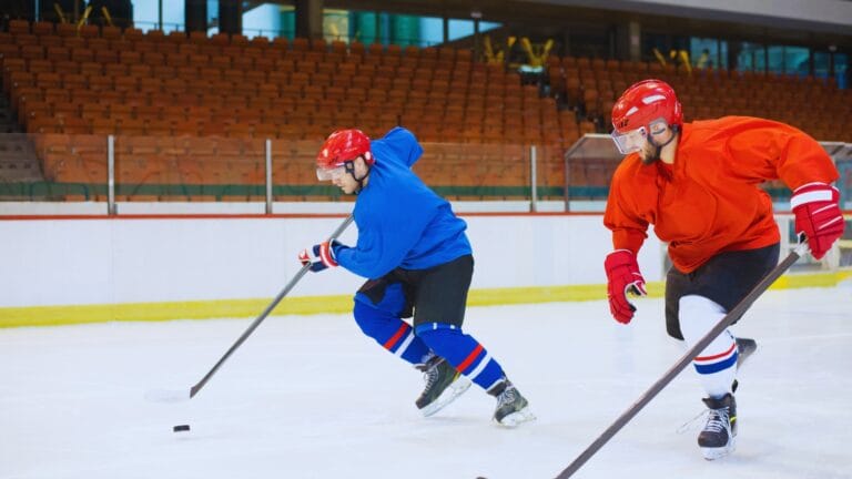 لقطة عمل احترافية للاعبي هوكي جليد يرتديان قمصانًا بألوان الأزرق والبرتقالي، ينزلجان بقوة على الجليد في ملعب هوكي جليد مغلق. أحدهما يهدف بالعصا نحو القرص، مع خلفية لمدرجات فارغة ذات مقاعد برتقالية وحاجز زجاجي. النص المدمج على الجليد: ما هي رياضة الهوكي.